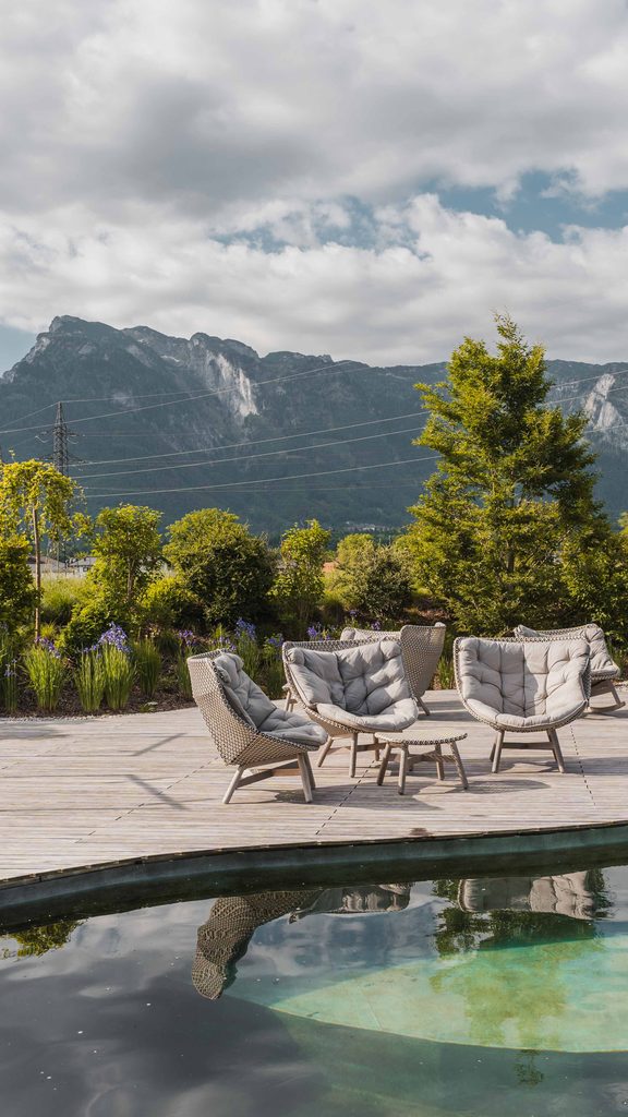 Loungesessel und Liegen auf Holzdeck am Naturpool mit Bergpanorama im Hintergrund.