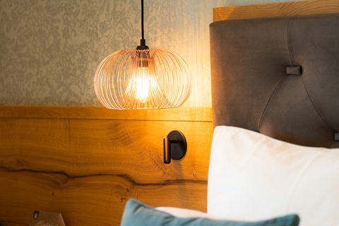 Detail eines Hotelbetts mit gepolstertem Kopfteil, Holzpaneel und leuchtender Hängelampe neben dem Nachttisch.