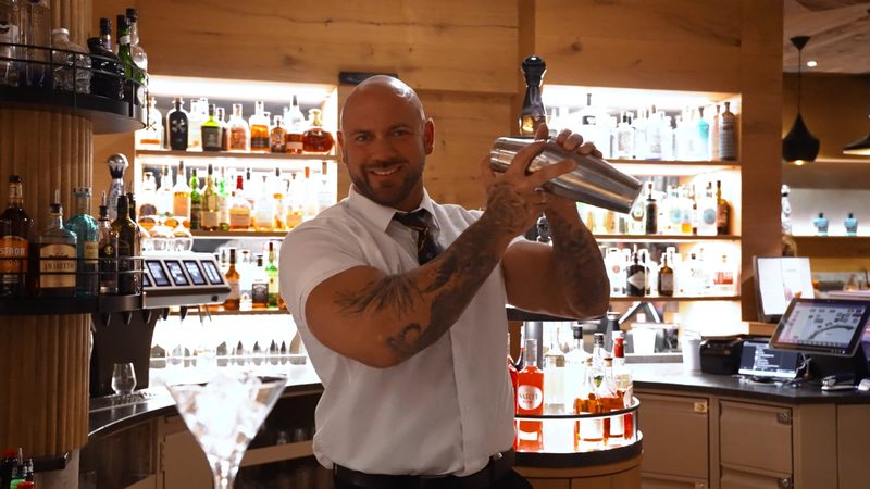 Barkeeper schüttelt einen Cocktailshaker an einer Hotelbar vor einem Regal mit Spirituosen.