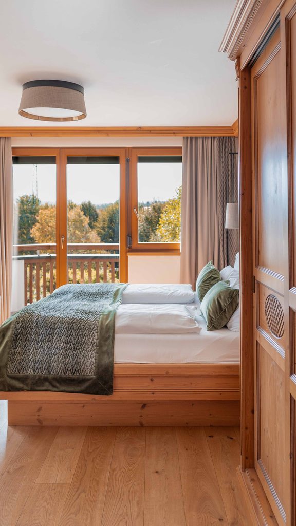 Holzdoppelbett in einem Hotelzimmer vor großer Fensterfront mit Balkon und Blick auf Bäume.