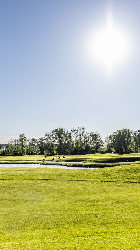 Wide golf course with pond, green fairways and trees under a clear sky.