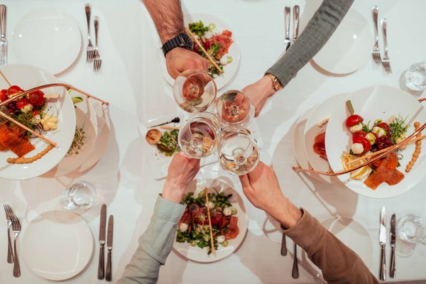 Mehrere Gäste stoßen mit Weingläsern über einem gedeckten Restauranttisch mit angerichteten Speisen an.