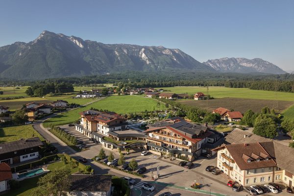 Luftaufnahme einer Hotelanlage mit Parkplatz vor grünen Wiesen und einem Alpenmassiv im Hintergrund.
